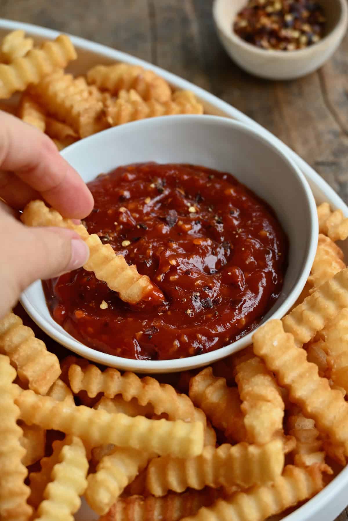 A hand is dipping a crinkle-cut French fry into a small white bowl of spicy ketchup that's nested into a larger white bowl of fries.