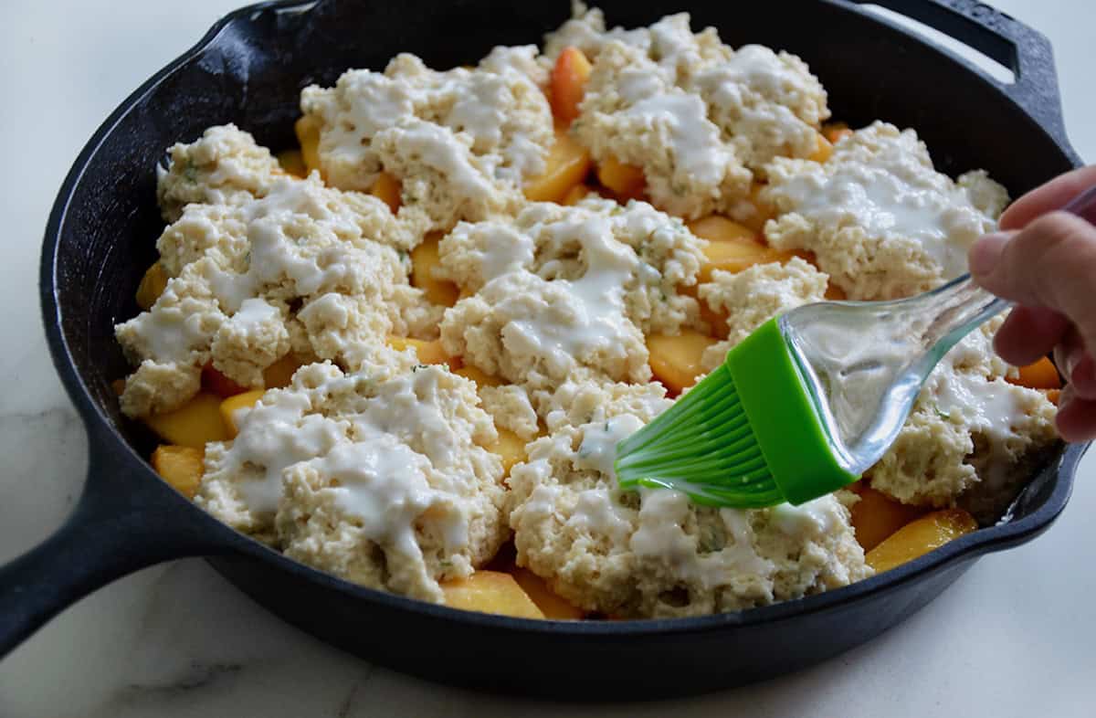 With a green silicone brush, buttermilk is being brushed onto the biscuits topping peach cobbler in a cast iron skillet.