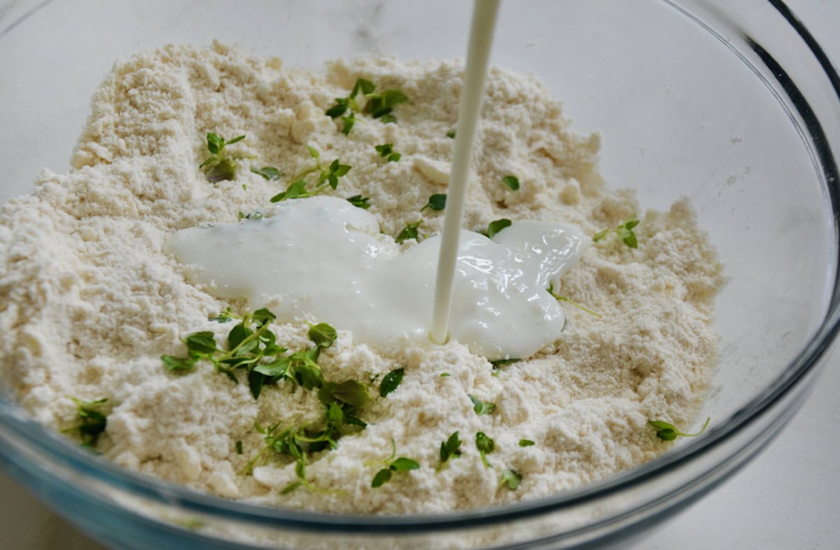 Buttermilk is being poured into a glass bowl filled with flour, fresh thyme and other biscuit ingredients.