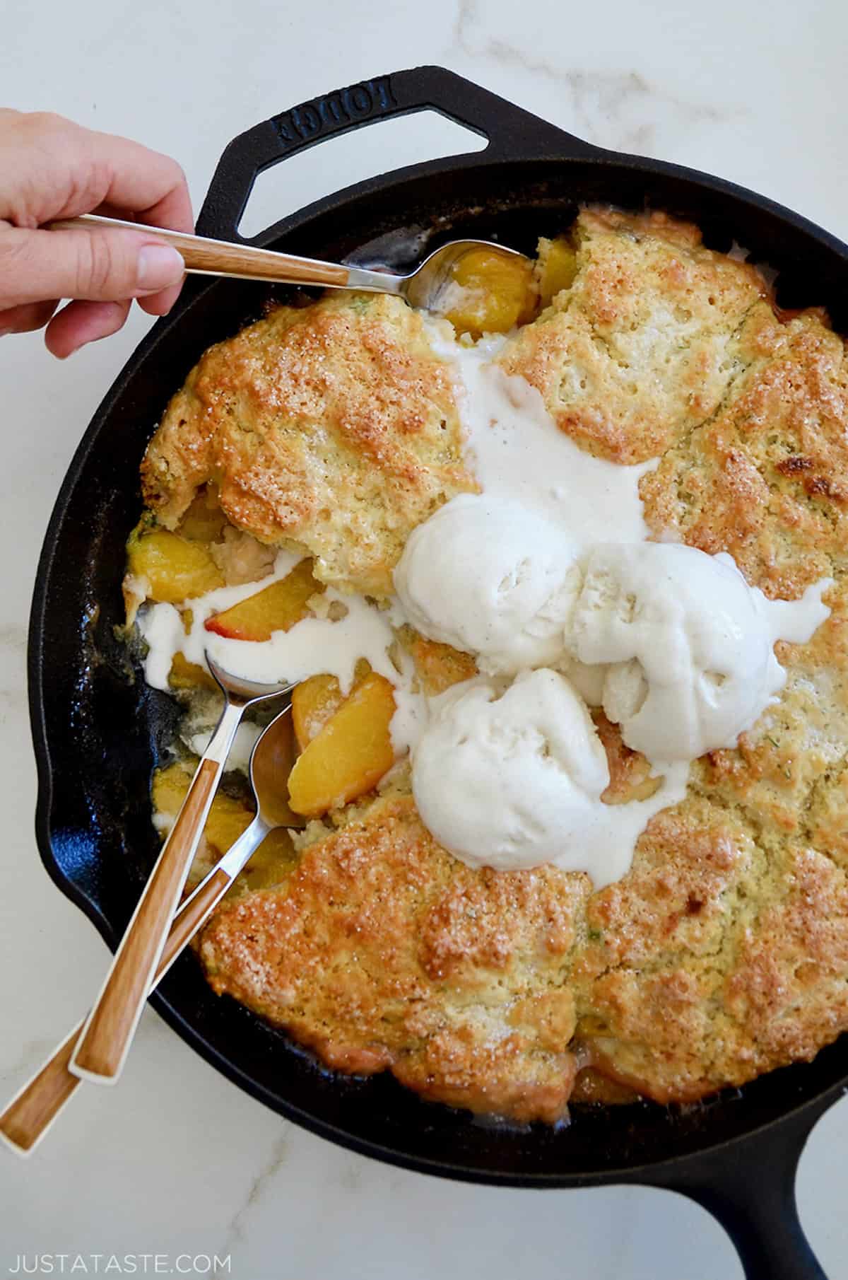 A hand is spooning up peach cobbler topped with buttermilk biscuit in a cast iron skillet. Two wood-handled spoons stick out of the cobbler.