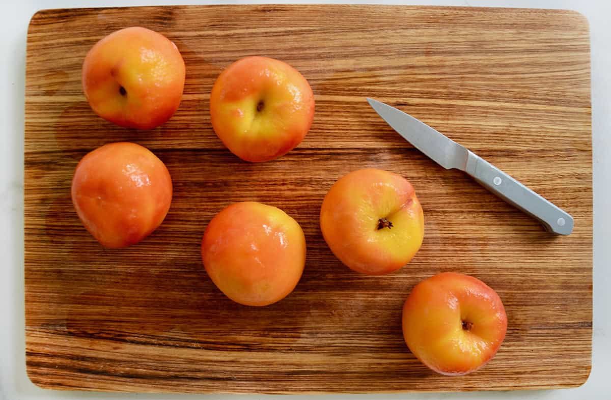 Peeled whole peaches are arranged on a wooden board, with a paring knife nearby.