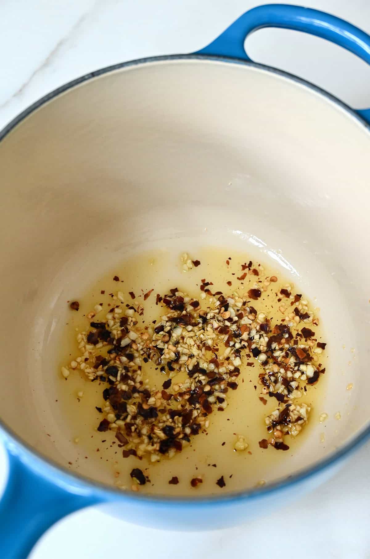 A blue enamel cast iron saucepan holds garlic and red pepper flakes being sauteed in oil.