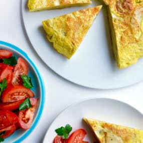 A golden-brown Spanish tortilla sits on a white plate. A wedge has been cut out and is on a separate plate along with a tomato salad. A blue bowl containing more tomato salad sits nearby.