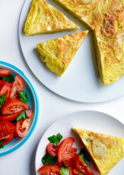A golden-brown Spanish tortilla sits on a white plate. A wedge has been cut out and is on a separate plate along with a tomato salad. A blue bowl containing more tomato salad sits nearby.