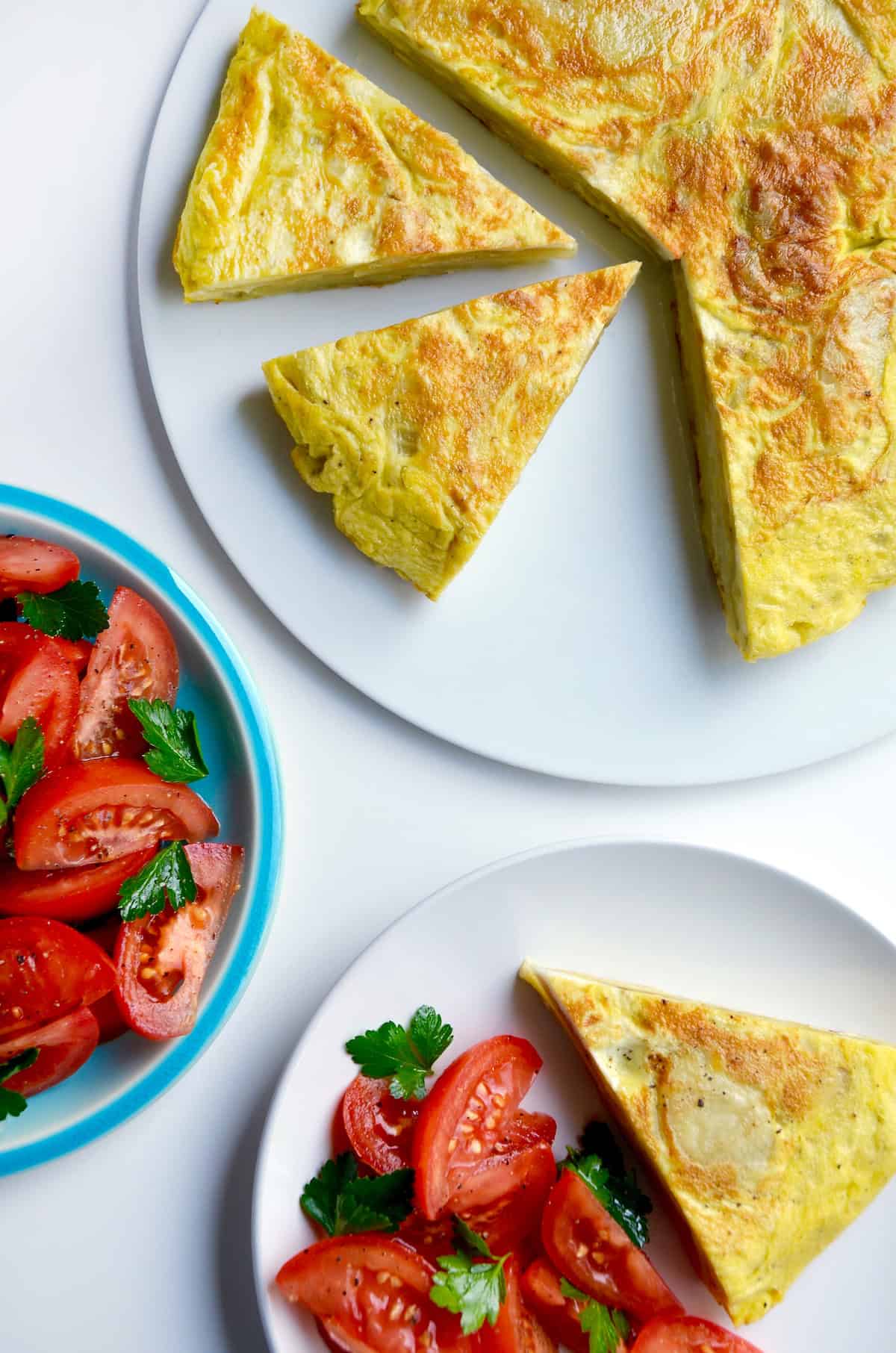 A golden-brown Spanish tortilla sits on a white plate. A wedge has been cut out and is on a separate plate along with a tomato salad. A blue bowl containing more tomato salad sits nearby.
