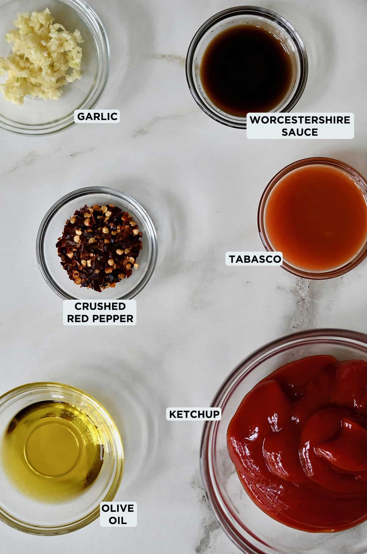 Small glass bowls on a white marble counter hold all the ingredients for spicy ketchup, including ketchup, olive oil, minced garlic, crushed red pepper and Tabasco.