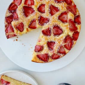 A strawberry-topped coffee cake sits on a white plate. One slice has been removed and is on a smaller plate with a fork. Nearby is a small sieve with powdered sugar.