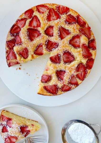 A strawberry-topped coffee cake sits on a white plate. One slice has been removed and is on a smaller plate with a fork. Nearby is a small sieve with powdered sugar.