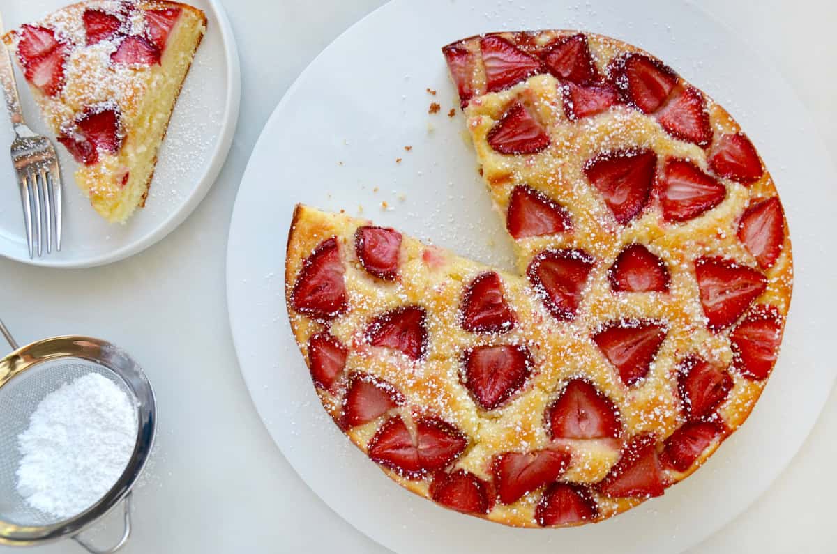 A strawberry-topped coffee cake sits on a white plate. One slice has been removed and is on a smaller plate with a fork. Nearby is a small sieve with powdered sugar.