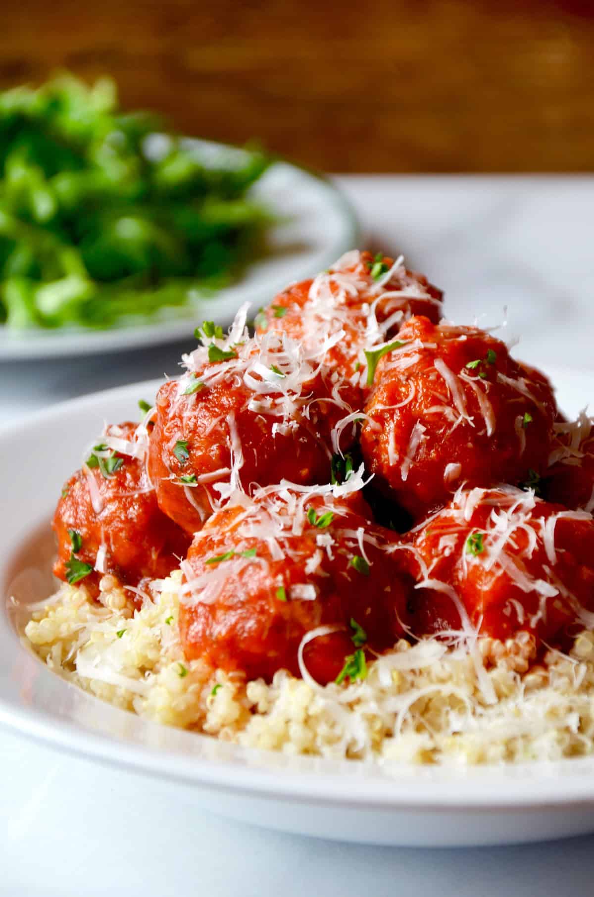 Baked turkey meatballs are coated in tomato sauce and sprinkled with parmesan cheese. They sit on a bed of quinoa in a white bowl. A white dish of greens is in the background.