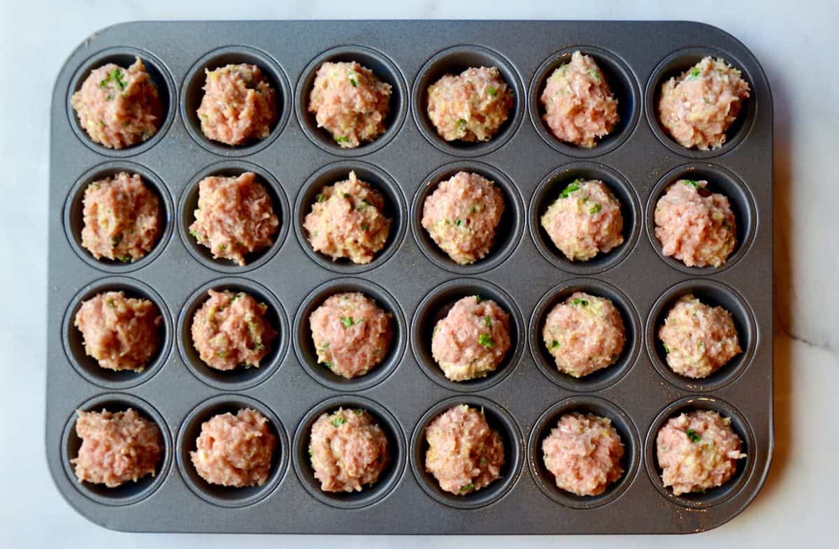 A mini muffin tin holds a raw turkey meatball in each cup.