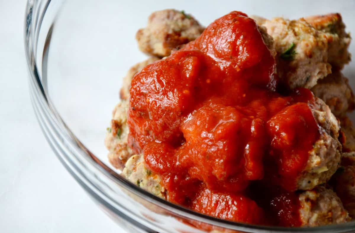 A clear glass bowl holds baked turkey meatballs with tomato sauce poured over them.