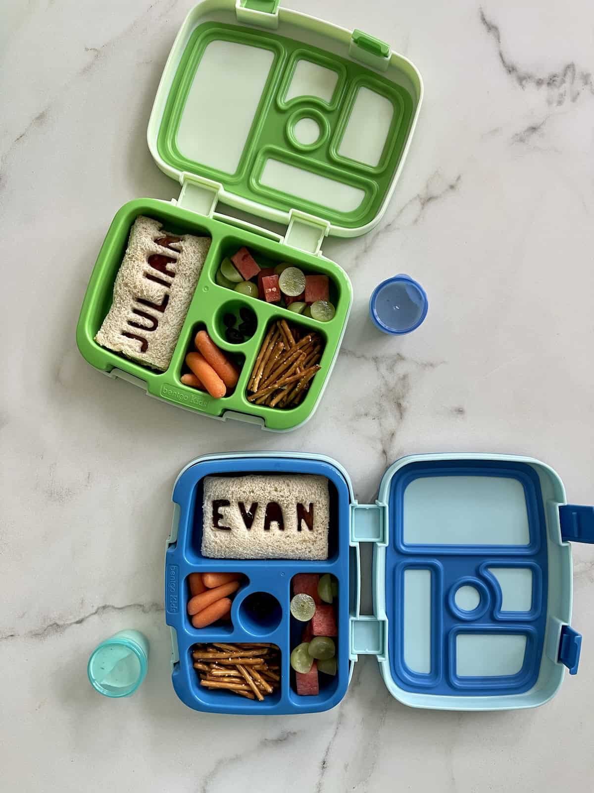 Bentgo lunchboxes, one green and one blue, with sandwiches, baby carrots, pretzel sticks and fresh fruit.