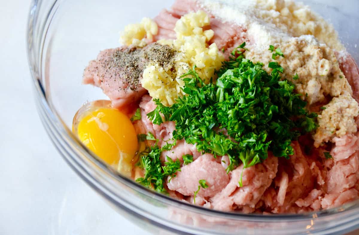 The ingredients for turkey meatballs are piled in a glass bowl, including ground turkey, egg, fresh parsley, breadcrumbs, minced garlic and seasonings.