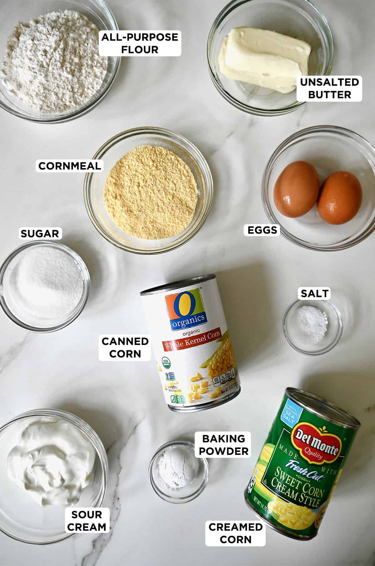 Corn casserole ingredients in various sizes of glass bowls along with canned creamed corn and canned whole kernel corn.