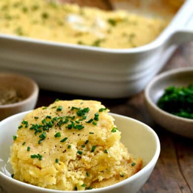 A scoop of corn casserole topped with fresh chives and melted butter in a small bowl.