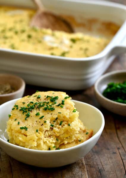 A scoop of corn casserole topped with fresh chives and melted butter in a small bowl.