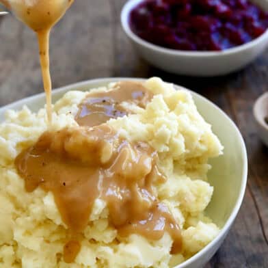 Homemade gravy being poured over mashed potatoes in a large serving bowl.
