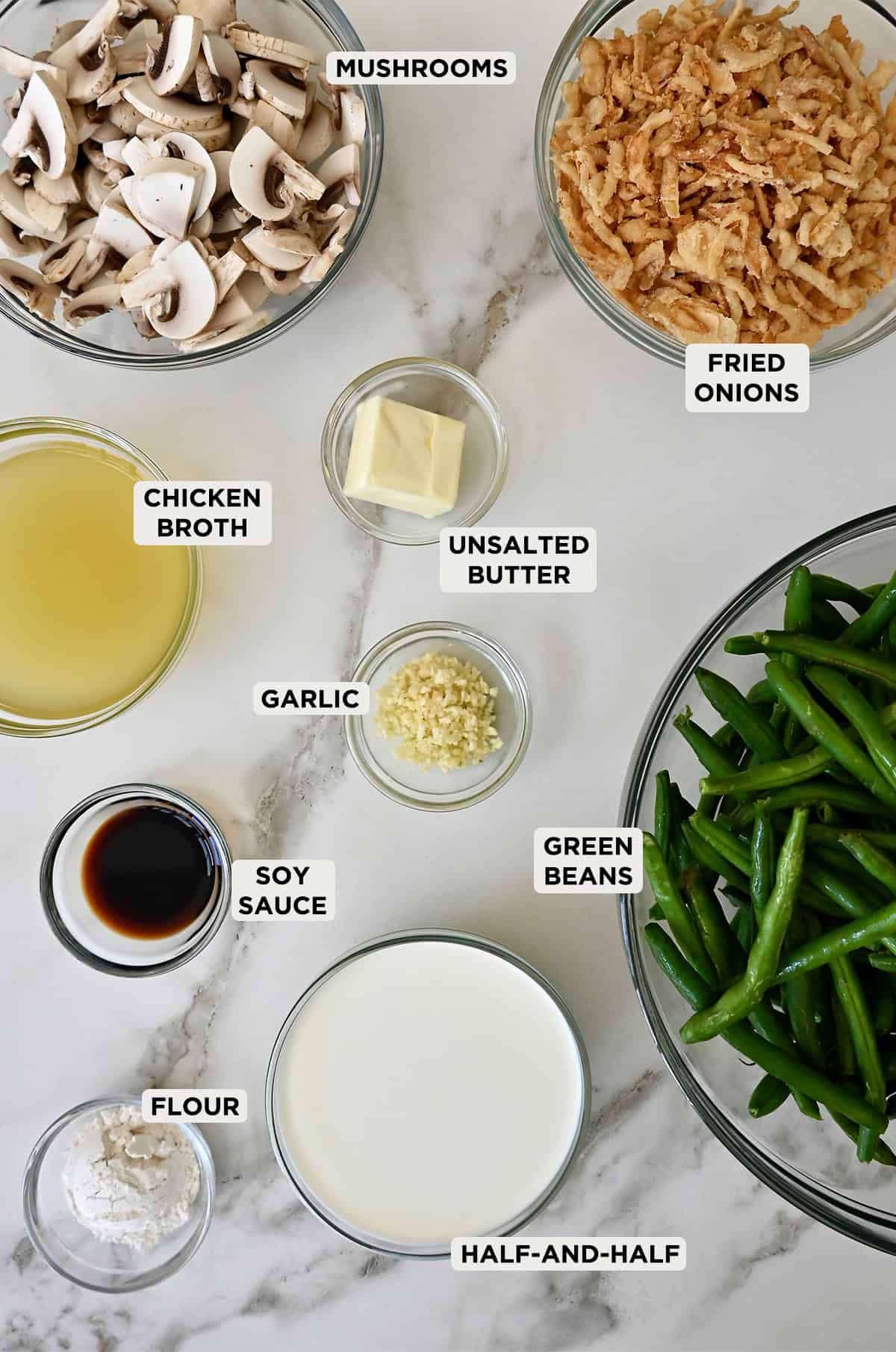 Green bean casserole ingredients in glass bowls, including fresh green beans, flour, half-and-half, mushrooms, butter, chicken broth, soy sauce and crispy fried onions.