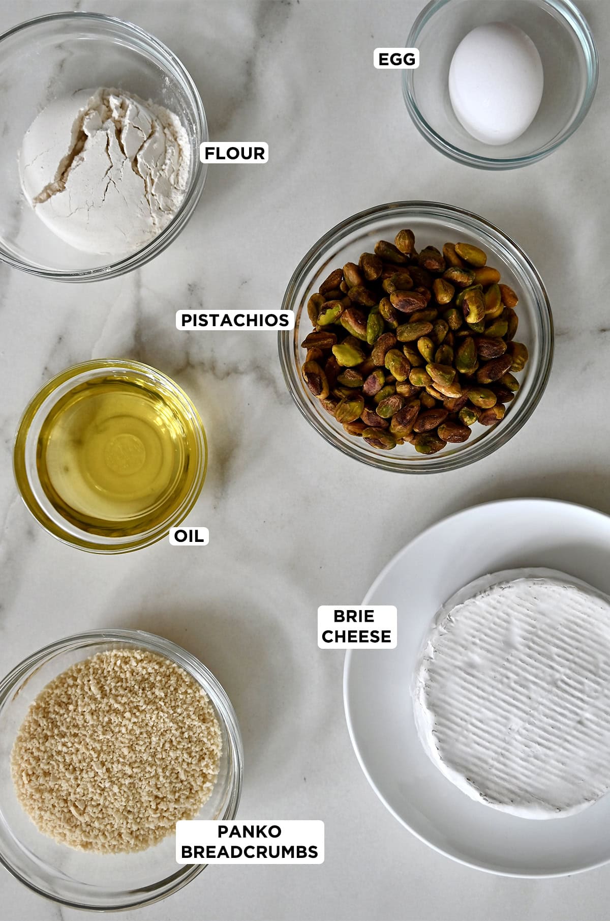 A wheel of brie cheese next to glass bowls containing pistachios, Panko breadcrumbs, oil, an egg and flour.