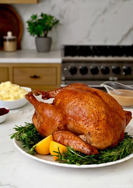 Roasted turkey on a serving plate surrounded by fresh thyme and a lemon wedge. Bowls of traditional Thanksgiving sides are nearby.