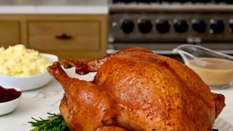 Roasted turkey on a serving plate surrounded by fresh thyme and a lemon wedge. Bowls of traditional Thanksgiving sides are nearby.