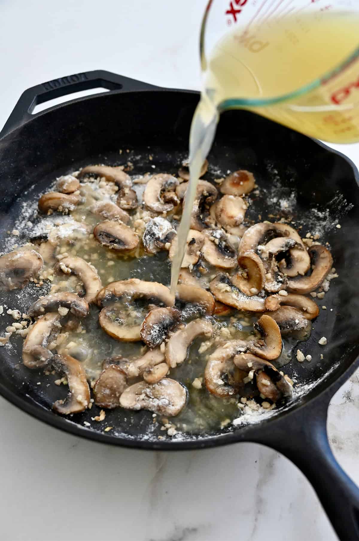 Chicken broth being added to a a skillet with sautéed mushrooms and garlic.