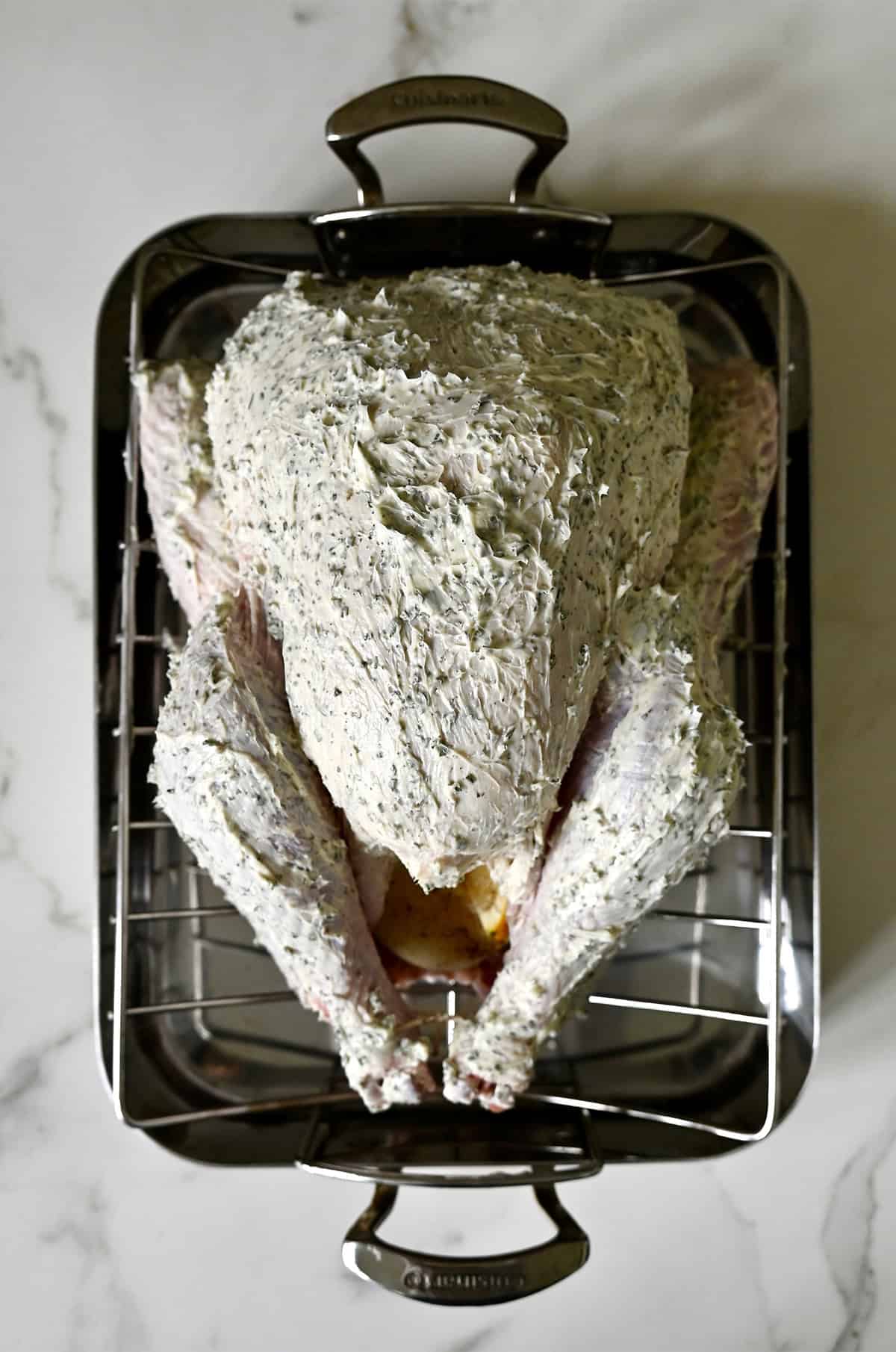 An uncooked turkey coated in herb butter atop a roasting rack inside of a roasting pan.