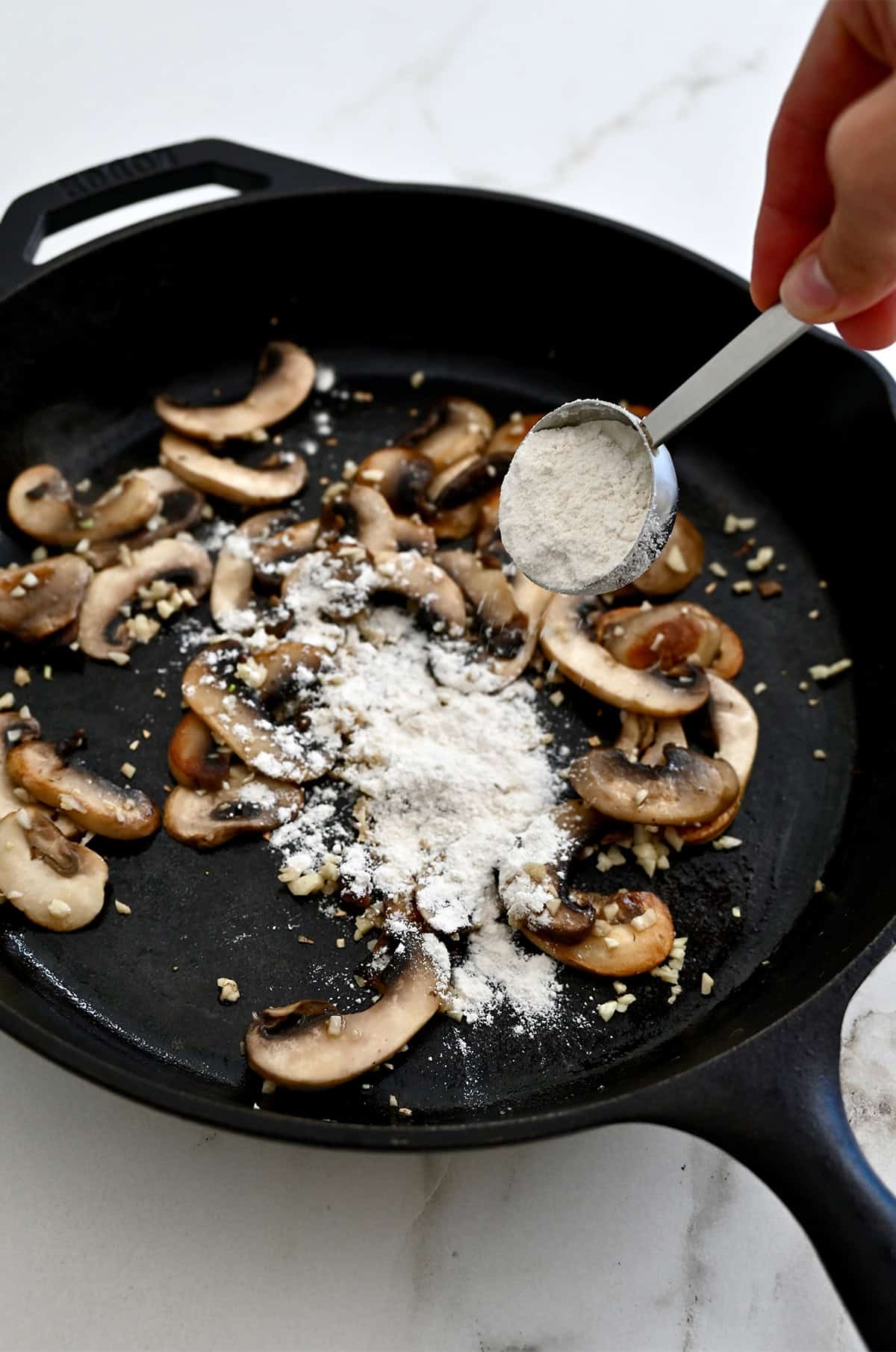 Flour being added to a skillet with sautéed mushrooms and garlic.