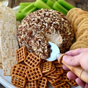 A classic cheese ball coated in chopped pecans on a plate surrounded by pretzels and crackers.