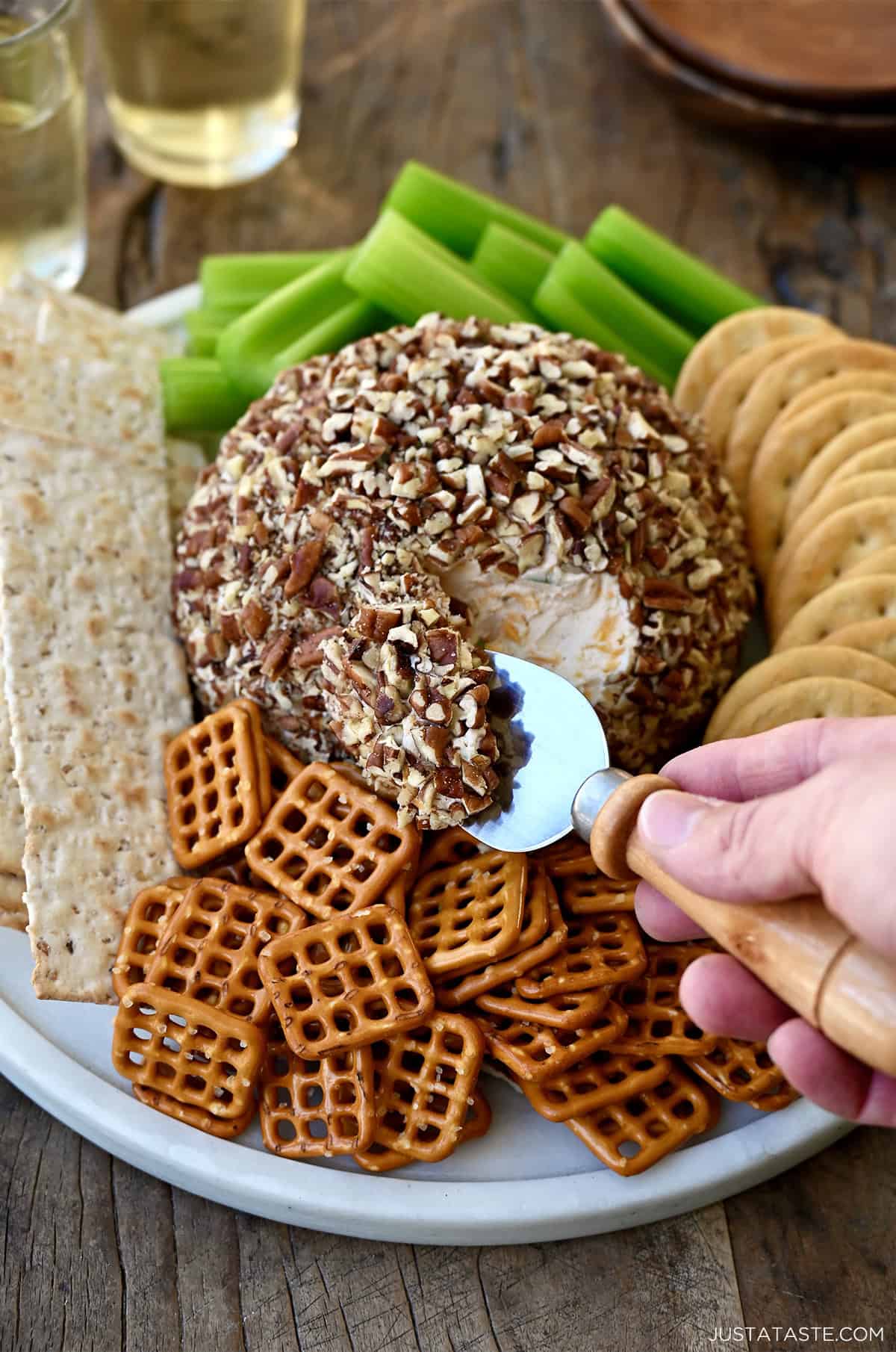 A classic cheese ball coated in chopped pecans on a plate surrounded by pretzels and crackers.