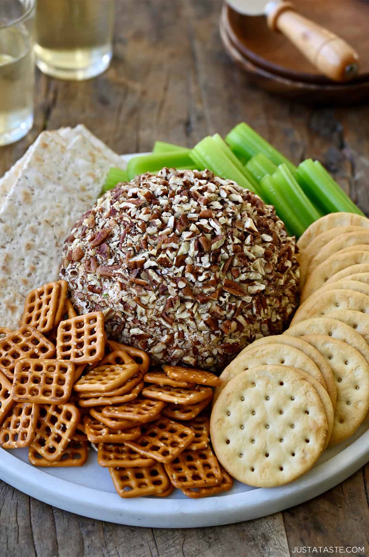 Cheese ball coated in crushed pecans on a serving plate with mini pretzels, crackers and celery sticks.