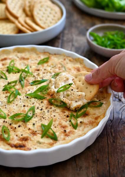 A hand dips a cracker into hot crab dip in a shallow serving dish.