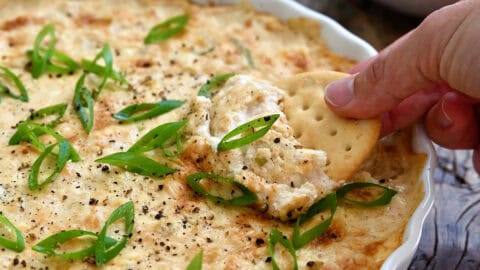 A hand dips a cracker into hot crab dip in a shallow serving dish.