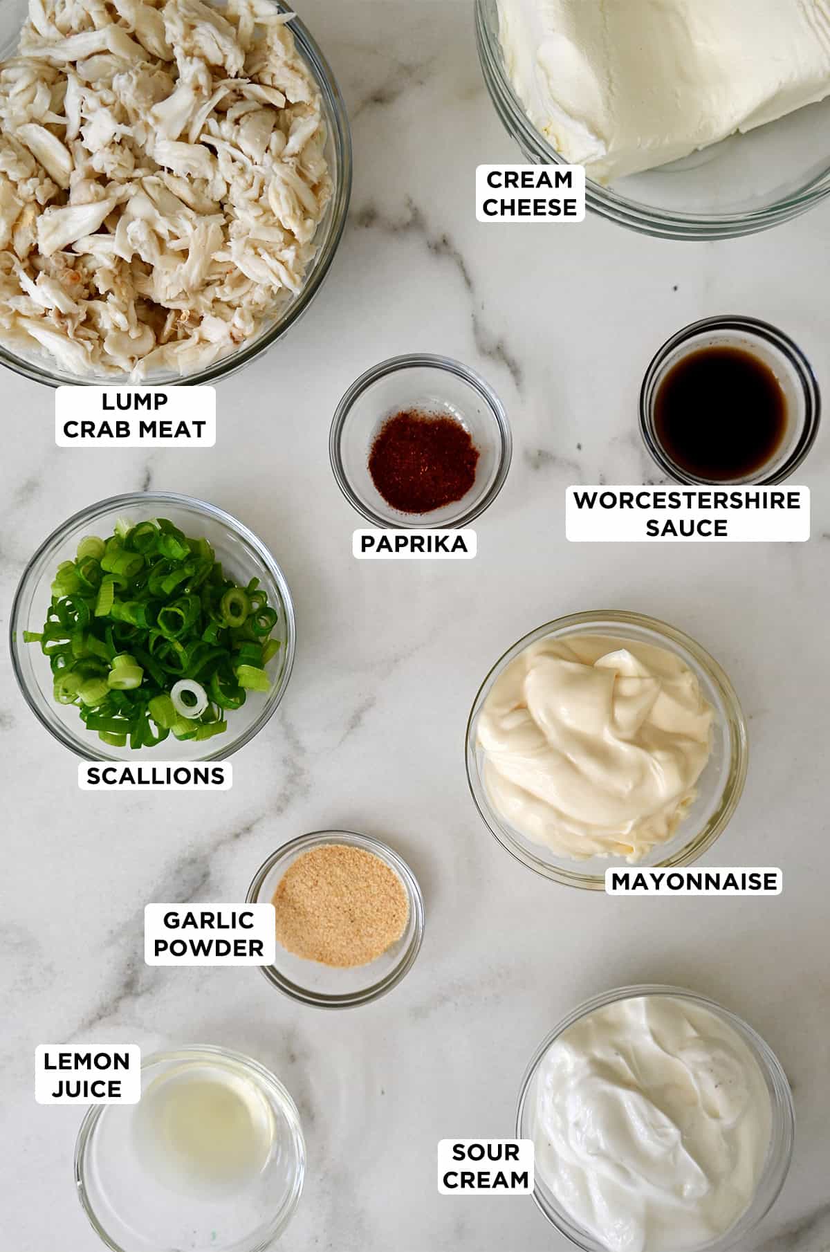 Maryland crab dip ingredients in glass bowls, including lump crab meat, mayonnaise, sour cream, scallions, Worcestershire sauce, paprika, cream cheese and lemon juice.