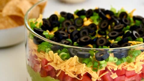 A dish containing 7 layer dip made with layers of refried beans mixed with taco seasoning, sour cream mixed with lime juice, guacamole, diced tomatoes, shredded cheddar cheese, scallions, and black olives.