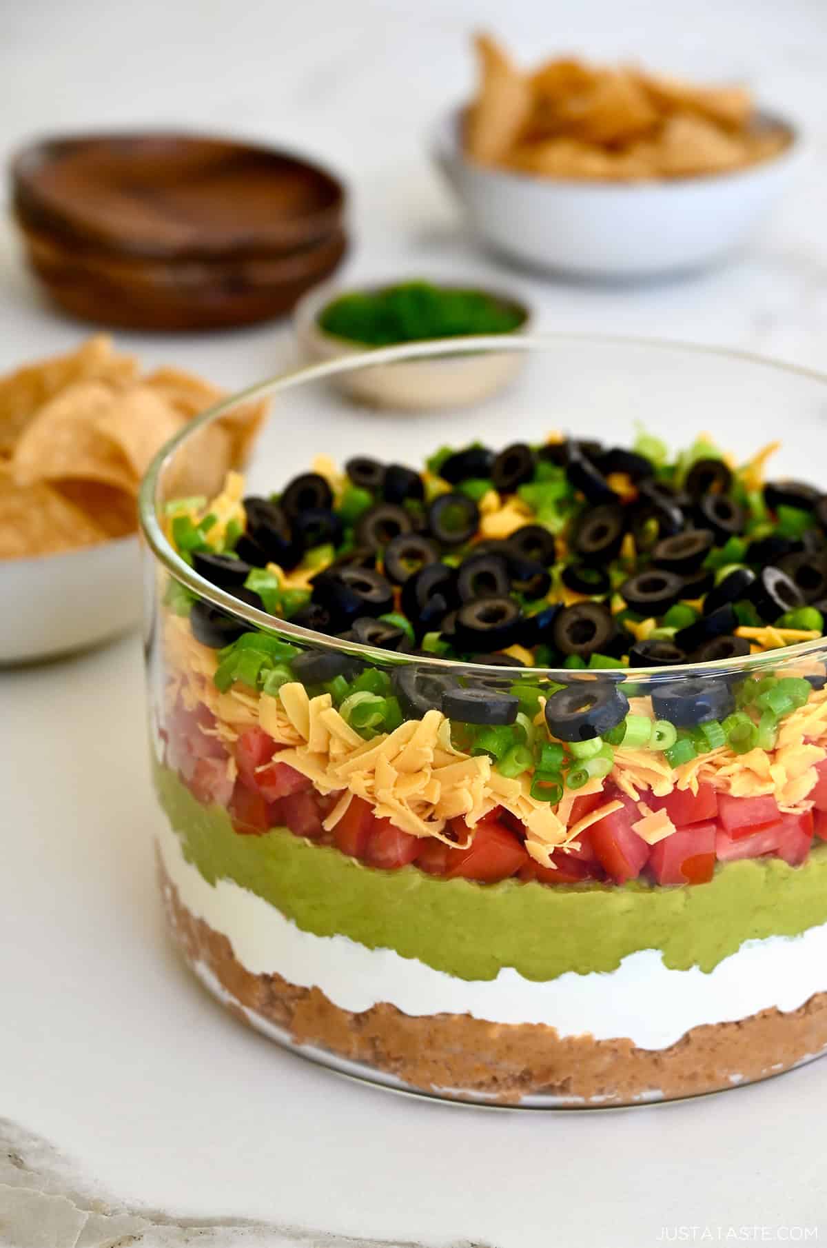 A dish containing 7 layer dip made with layers of refried beans mixed with taco seasoning, sour cream mixed with lime juice, guacamole, diced tomatoes, shredded cheddar cheese, scallions, and black olives.