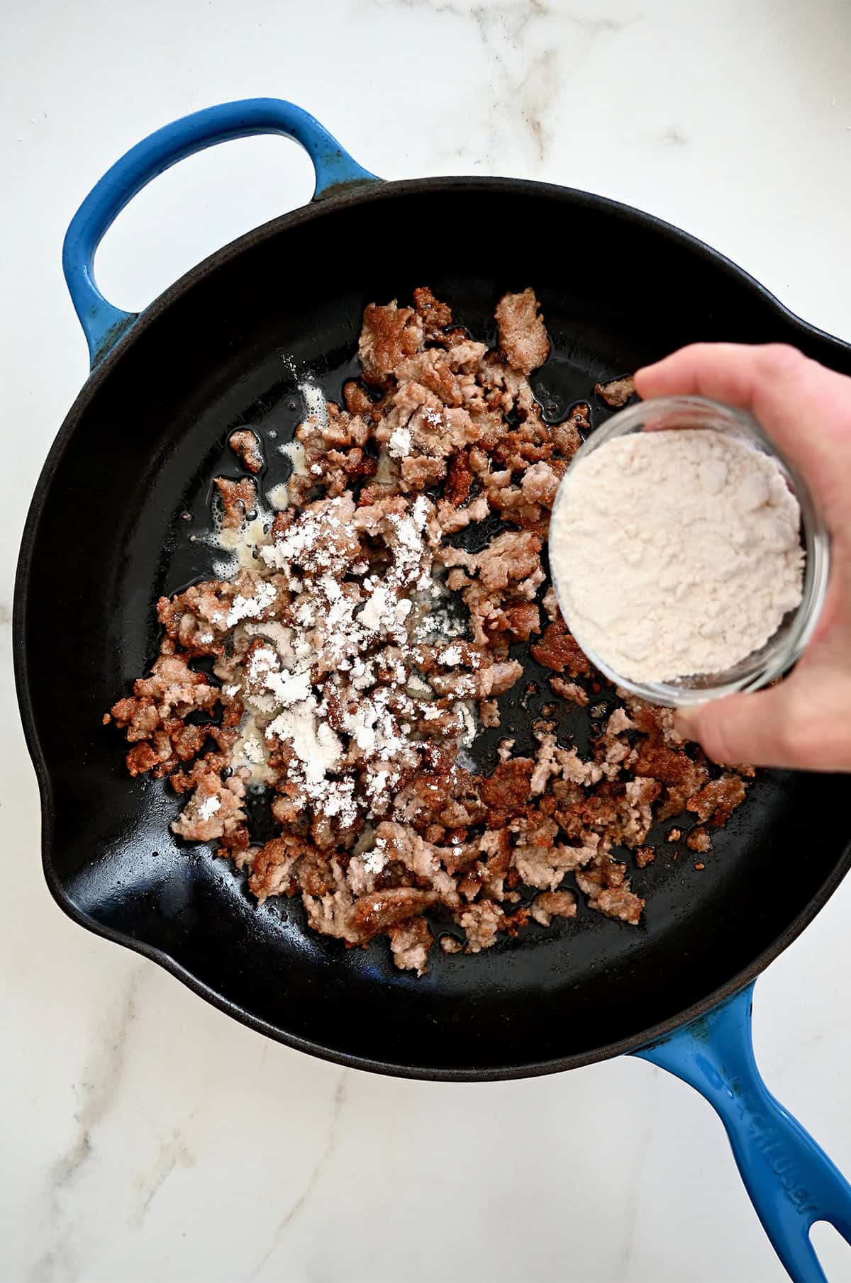 Adding flour atop browned pork sausage in a skillet.