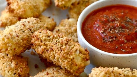 Homemade air fryer mozzarella sticks on a plate with a small bowl containing marinara sauce.
