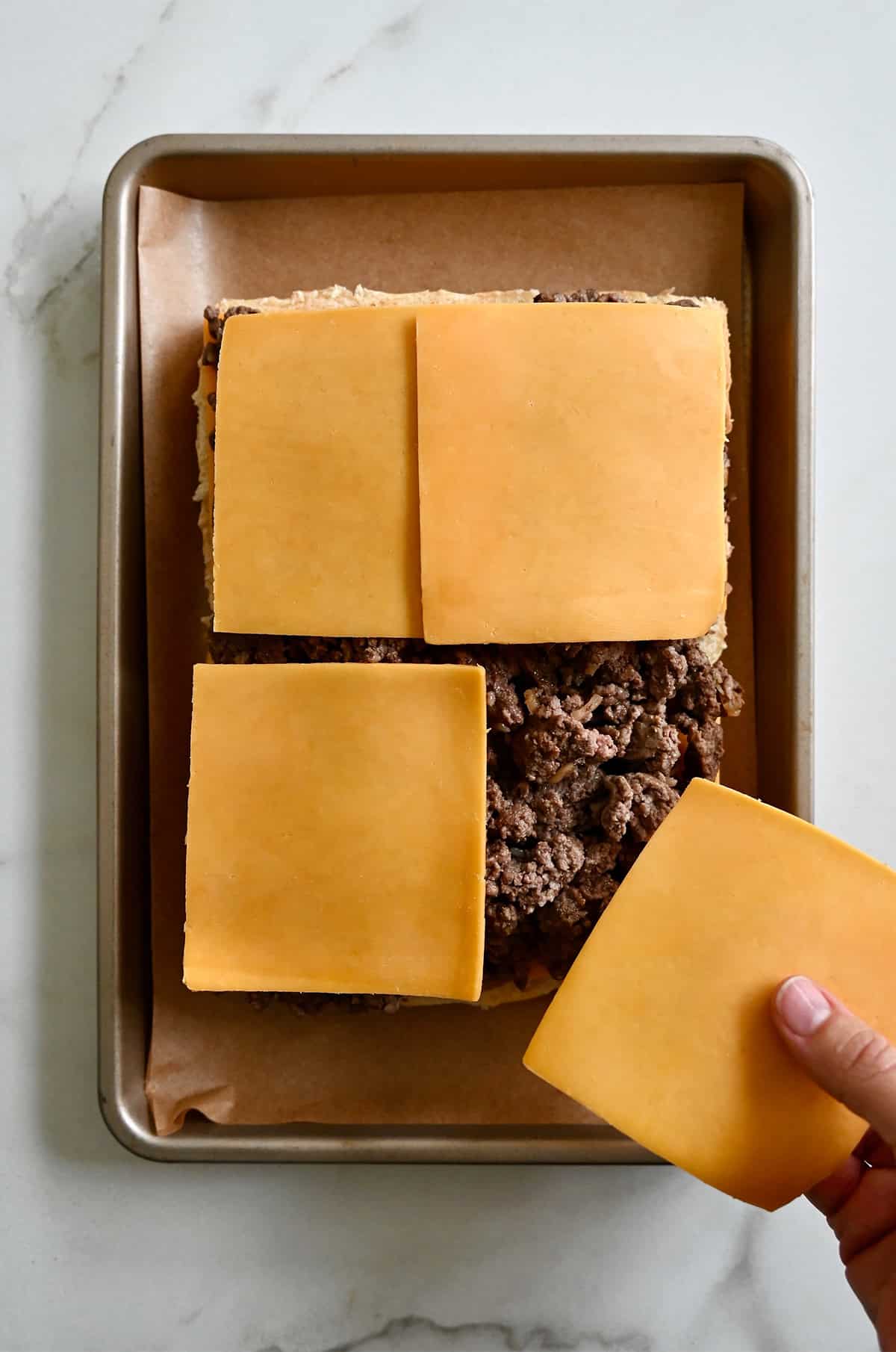 Buns topped with cooked burger meat being topped with slices of cheese.