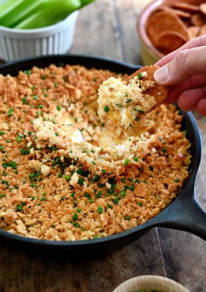 A hand holding a Ritz cracker dips it into a cheesy jalapeño popper dip in a cast iron skillet.