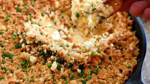 A hand holding a Ritz cracker dips it into a cheesy jalapeño popper dip in a cast iron skillet.