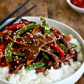 Beef stir-fry with veggies atop steamed white rice on a plate with chopsticks.