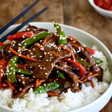 Beef stir-fry with veggies atop steamed white rice on a plate with chopsticks.