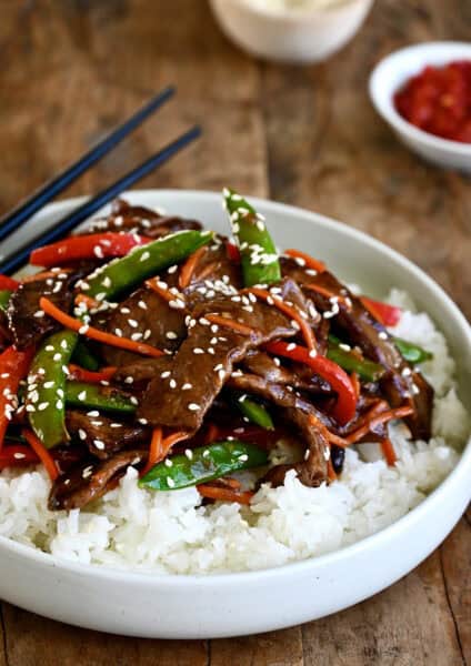 Beef stir-fry with veggies atop steamed white rice on a plate with chopsticks.