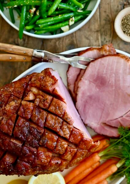 Baked ham with brown sugar glaze on a serving plate with roasted veggies.