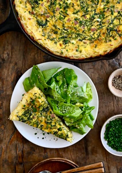 A slice of ham and cheese quiche with a crispy hash brown crust on a plate with dressed greens.