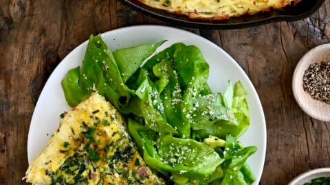 A slice of ham and cheese quiche with a crispy hash brown crust on a plate with dressed greens.