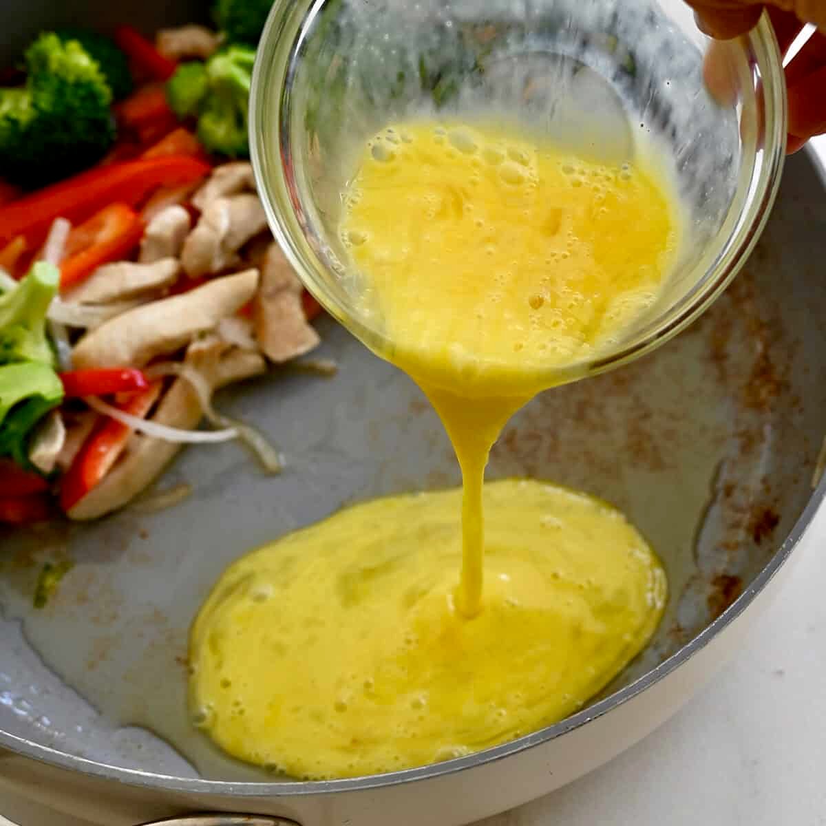 Adding whisked eggs to a hot skillet with stir-fried chicken and veggies.