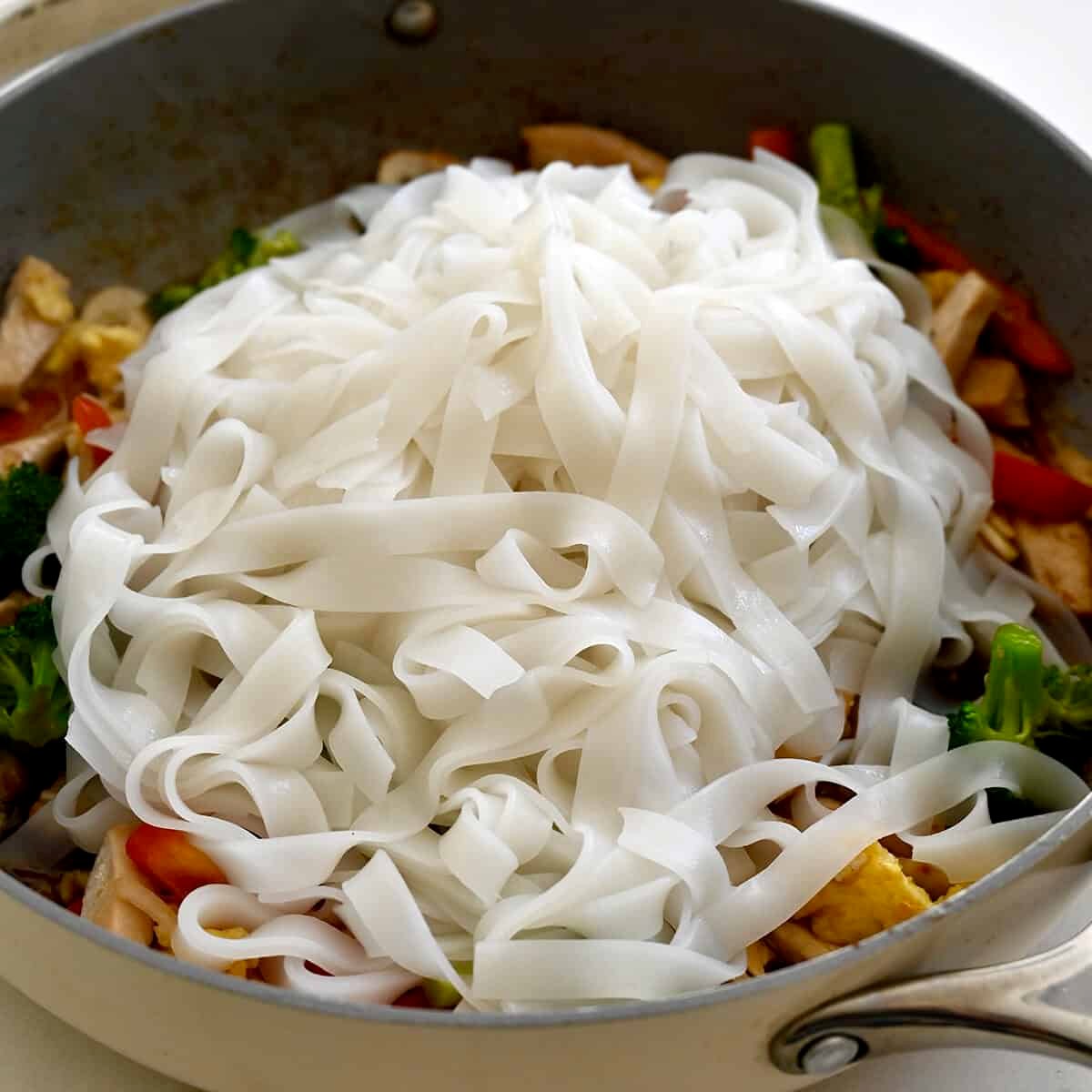 Cooked rice noodles atop stir-fried chicken and veggies in a hot skillet.